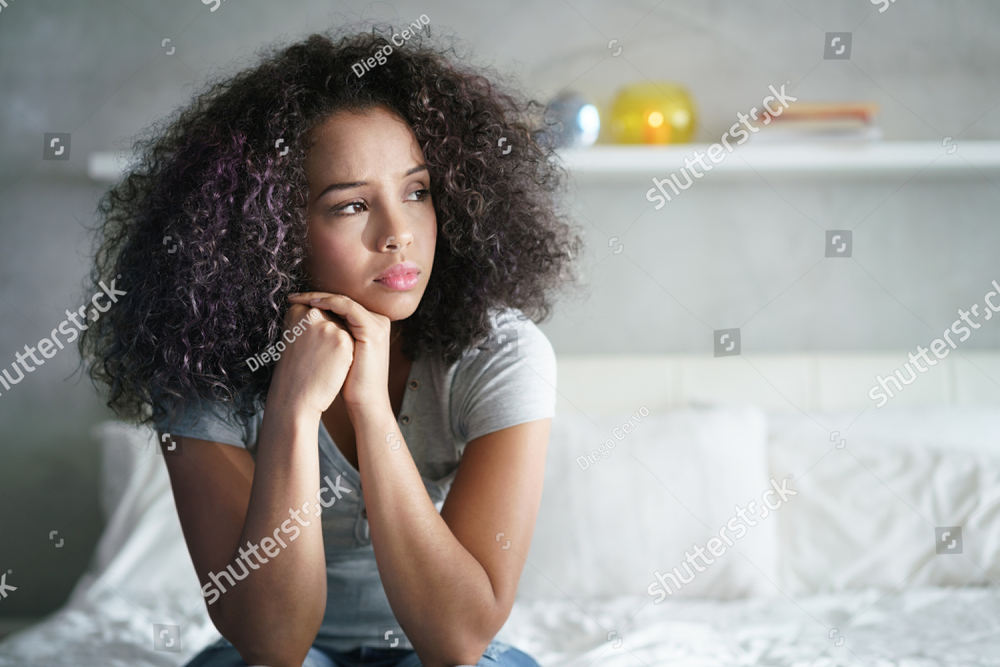 A teenage girl sitting on her bed, chin resting on her hands, looking away from the camera.