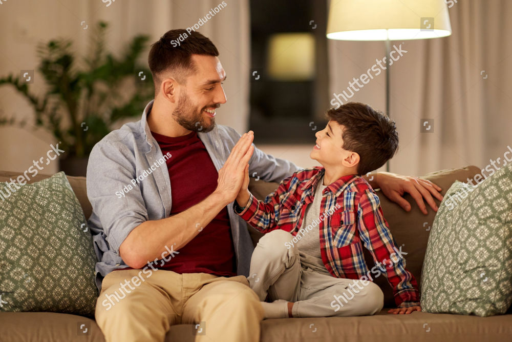 A father and his young son sitting on a sofa, mid high-five, both laughing.