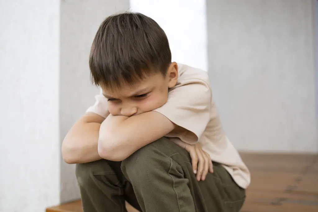 A young boy sitting on the floor with his arms folded over his knees, head down.