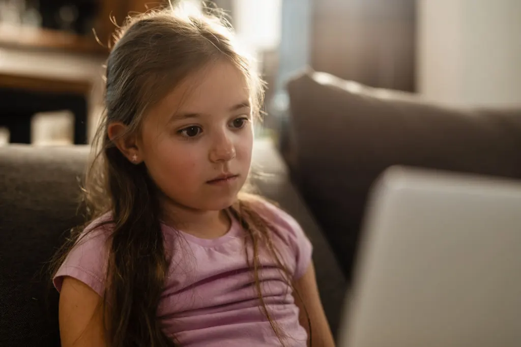 A young girl sitting on a sofa looking at a laptop, thoughtful.